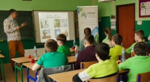 Na zdjęciu Doradca Energetyczny stojący przy wyświetlaczu multimedialnym omawiający sposoby produkcji energii. W tle dzieci w wieku szkolnym siedzące i słuchające Doradcę.