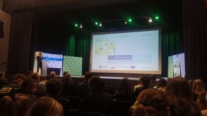 Zdjęcie przedstawiające salę konferencyjna, w której siedzą uczestnicy konferencji. Na podeście znajduje się Doradca energetyczny prezentujący slajdy. 