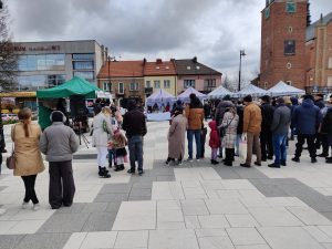 Zdjęcie przedstawiające grupę ludzi i dzieci stojących na rynku. W tle namiotu wystawiennicze.